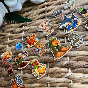 May include: A collection of colorful, cartoonish stickers featuring various characters and objects. Designs include a shark on a surfboard, a teacup ballerina, a monkey in a banana, and a creature with the text "Tung Tung Tung Sahur". The stickers are arranged on a woven surface.