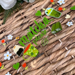 May include: A green and white sign with the text "Matcha Please" in cursive. The sign is decorated with green hearts, flowers, and a teacup, tea bag, and boba tea.