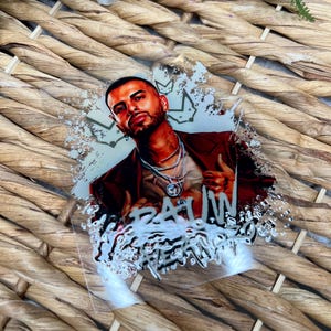 May include: A close-up of a sticker featuring a portrait of a man with a crown and the word "RAW" in white lettering. The sticker has a white and gray splatter effect.