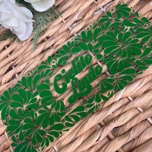 May include: A green acrylic bookmark with the words "Cielito Lindo" in cursive script, surrounded by a floral design. The bookmark is set against a woven wicker background, with white flowers in the upper left corner.