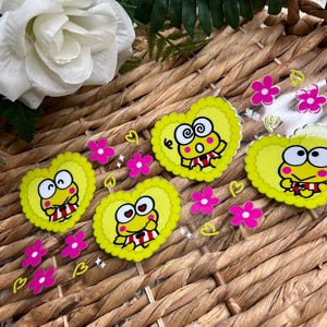 May include: Four yellow heart-shaped stickers with a cartoon frog wearing a red and white striped shirt. The stickers are on a clear plastic sheet with pink flowers and hearts.