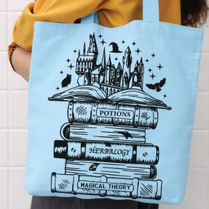 May include: A light blue tote bag featuring a black graphic print of a stack of books titled Potions, Herbalogy, and Magical Theory. An open book sits atop the stack, with a castle, a witch hat, stars, and two owls illustrated above it.