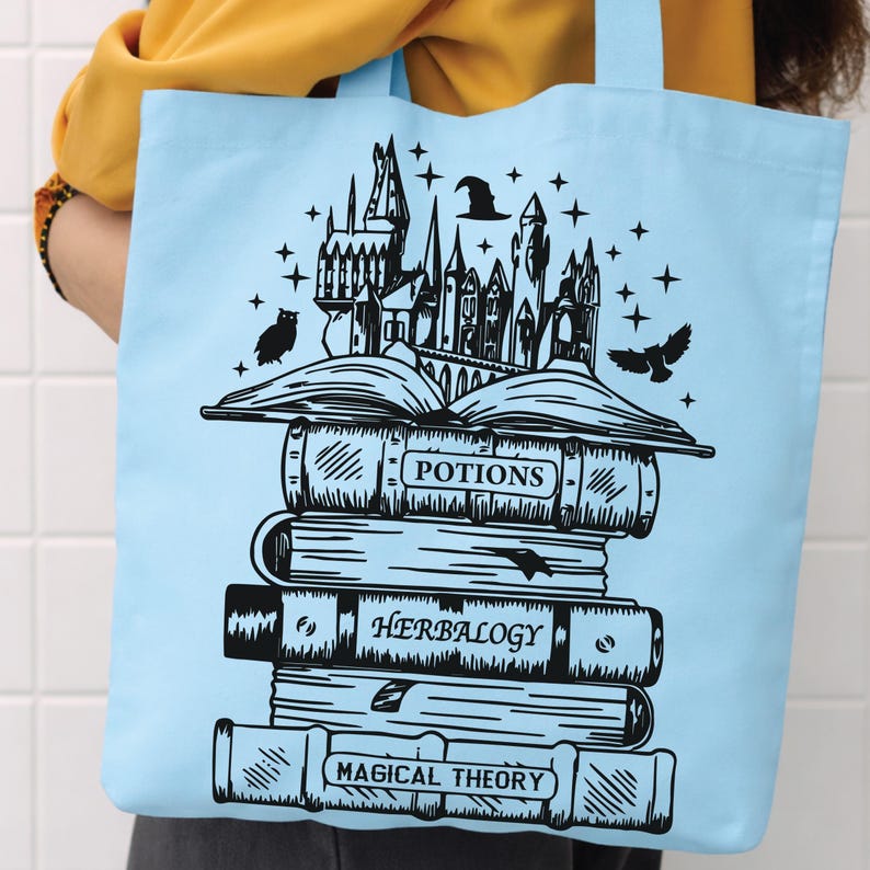 May include: A light blue tote bag featuring a black graphic print of a stack of books titled Potions, Herbalogy, and Magical Theory. An open book sits atop the stack, with a castle, a witch hat, stars, and two owls illustrated above it.
