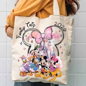 May include: Cream-coloured tote bag with a vibrant graphic of Disney characters, including Mickey and Minnie Mouse, Goofy, Donald and Daisy Duck, and Pluto. The words "Family Trip 2026" are printed on the bag, along with a castle and fireworks.