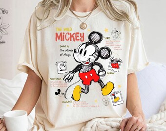 Peut inclure: Un t-shirt couleur crème avec un graphisme style croquis de Mickey Mouse en noir, rouge et jaune. Le texte indique THE WILD MICKEY, Exhibit A: The Mouse of Magic, ainsi que des sections intitulées STATS, SIGHTINGS, ENJOYS et FEATURES avec de petites images style polaroïd.