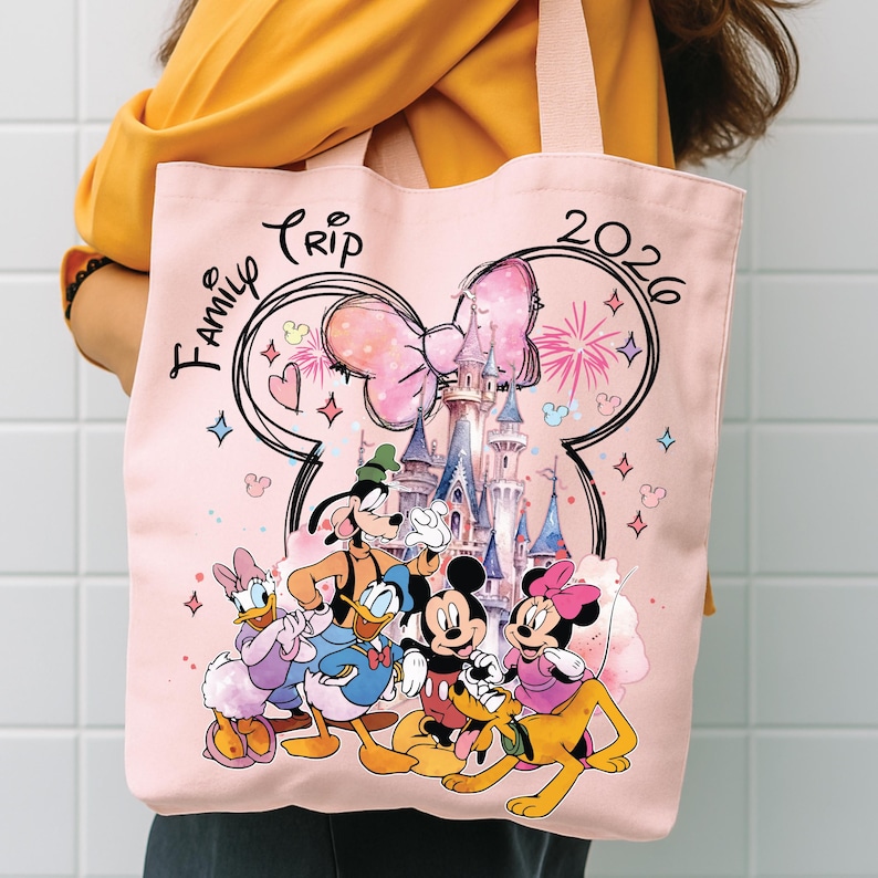 May include: A pink tote bag with a Disney design. The bag features Mickey Mouse, Minnie Mouse, Donald Duck, Daisy Duck, Goofy, and Pluto in front of a castle. The words "Family Trip 2026" are printed on the bag.