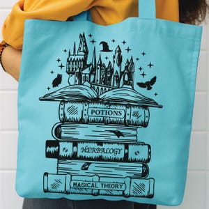 May include: A light blue tote bag featuring a black graphic print of a stack of books titled Potions, Herbalogy, and Magical Theory. An open book sits atop the stack, topped with a castle silhouette, a witch hat, stars, and two owls.