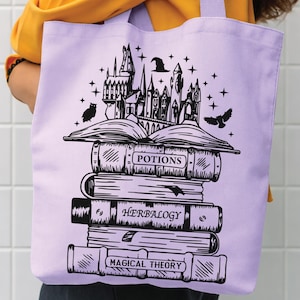 May include: A light purple tote bag featuring a black graphic print of a stack of books topped with a castle, an owl, and a witch hat. The book spines are labelled with the text Potions, Herbalogy, and Magical Theory in a clear, bold font.