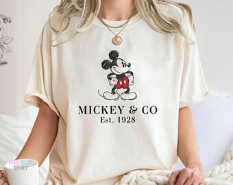 Peut inclure: Un t-shirt à manches courtes de couleur crème arborant un dessin au trait noir et blanc de Mickey Mouse avec un short rouge. Sous le motif, le texte MICKEY & CO et Est. 1928 est imprimé en police serif noire au centre de la poitrine.