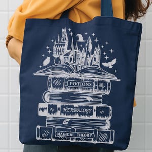 May include: A navy blue tote bag featuring a white graphic print of a stack of books labelled Potions, Herbalogy, and Magical Theory. An open book sits atop the stack, with a castle, a witch hat, stars, and two owls illustrated above it.