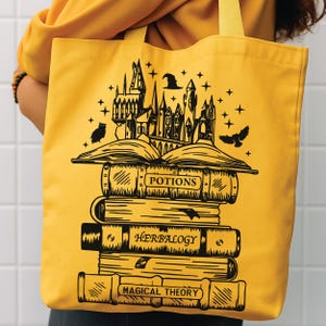 May include: A bright yellow tote bag featuring a black graphic print of a stack of books labelled Potions, Herbology, and Magical Theory. An open book sits atop the stack, with a castle, a witch hat, stars, and two flying owls above it.