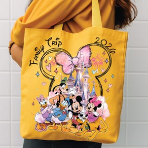 May include: Yellow tote bag with a Disney castle and characters, including Mickey and Minnie Mouse, Donald Duck, and Goofy. The bag has the words "Family Trip 2026" and a large pink bow design.