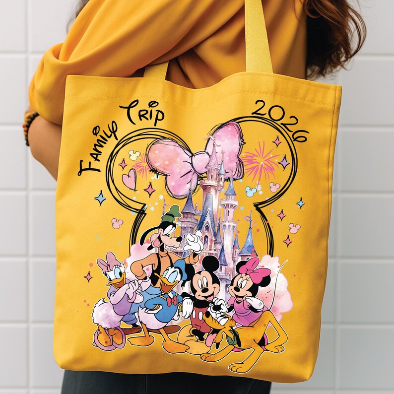 May include: Yellow tote bag with a Disney castle and characters, including Mickey and Minnie Mouse, Donald Duck, and Goofy. The bag has the words "Family Trip 2026" and a large pink bow design.