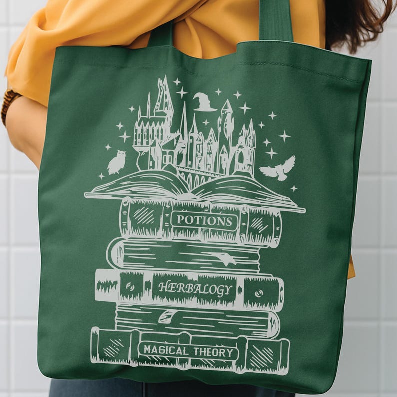 May include: A dark green tote bag featuring a white line art graphic of a stack of books topped with an open book and a castle silhouette. The book spines are labelled Potions, Herbalogy, and Magical Theory, with stars and a hat above.