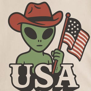 May include: Cream-colored t-shirt featuring a green alien wearing a red cowboy hat and holding an American flag. The word "USA" is printed in bold, white letters below the alien. The shirt is folded on a woven mat.