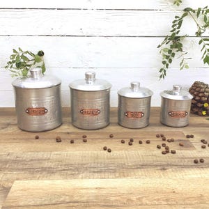 May include: Four vintage metal canisters with lids, labeled "SUCRE", "FARINE", "THE", and "SPICES" in copper-colored labels. The canisters are silver-toned and sit on a wooden surface. A pinecone and greenery are in the background.