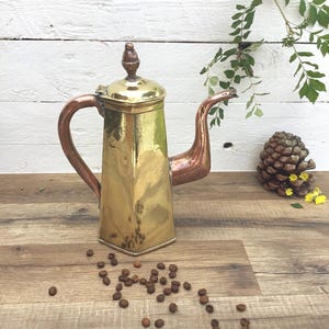 Puede incluir: Una cafetera vintage de latón con asa y boquilla de cobre. Tiene una base cuadrada y una tapa decorativa. Granos de café esparcidos sobre una superficie de madera. El fondo muestra una pared de madera blanca y vegetación.