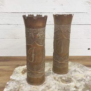 May include: Two antique brass vases with ornate embossed designs and a shield with the letter "J". The vases have a scalloped rim and are cylindrical in shape. They are displayed on a stone surface against a white wooden background.