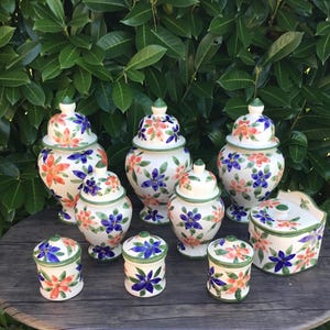 Peut inclure: Ensemble de pots et récipients en céramique avec couvercles, ornés de motifs floraux orange, bleu et vert. Les pots sont de tailles et de formes variées, avec une base blanche et une bordure verte, disposés sur une table en bois.