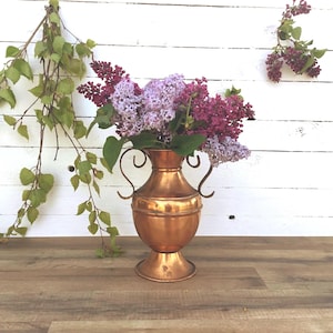 Könnte beinhalten: Eine Kupfervase mit zwei Henkeln, gefüllt mit einem Strauß lila und weißer Fliederblüten. Die Vase steht auf einer Holzoberfläche vor einer weißen Holzvertäfelung.