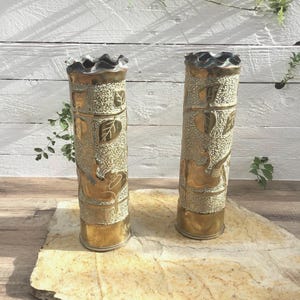 Puede incluir: Dos jarrones de latón con diseños de hojas y texturas. Los jarrones tienen un acabado dorado con una parte superior con volantes. Son cilíndricos y se asientan sobre una losa de piedra.
