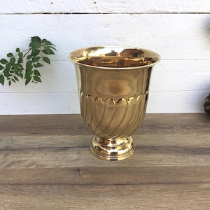 May include: A gold-colored metal urn with a wide rim and a pedestal base. The body of the urn features vertical, curved grooves. The urn is displayed on a wooden surface with a white background and greenery.