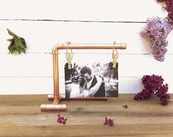 May include: Copper pipe photo display stand with two wooden clothespins holding a black and white photo of a couple. The stand is on a wooden surface.