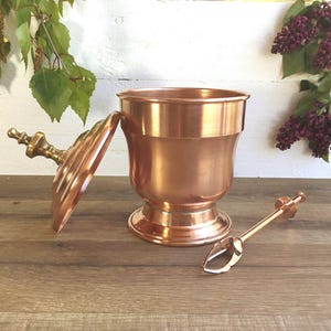 May include: A copper ice bucket with a lid and a matching copper ice tong. The bucket has a round, flared base and a smooth, polished finish.