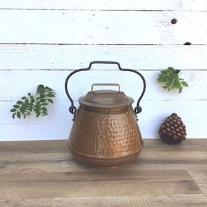 May include: A copper-colored, hammered metal pot with a lid and a black handle. The pot has a rounded shape and a small handle on the lid. The background is a white wooden wall and a wooden surface.
