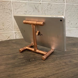 Peut inclure: Un support de tablette en tuyau de cuivre. Le support est fait de tuyaux et de raccords en cuivre, avec un support en forme de T pour la tablette. La tablette est argentée et repose sur le support. Le support est sur une surface en bois.