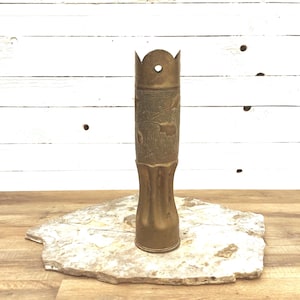 Puede incluir: Un jarrón de latón vintage con un diseño en relieve. El jarrón tiene una base acampanada y una parte superior en forma de corona con un agujero. Se exhibe sobre una losa de piedra natural contra un fondo de madera blanca. El jarrón mide unos 25 cm de alto.