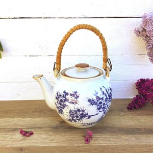 Peut inclure: Théière en céramique blanche avec un motif floral bleu. La théière a une anse tressée et un couvercle en bois. Le bec verseur est incurvé et elle est posée sur une surface en bois. Des fleurs décoratives sont en arrière-plan.