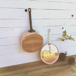 May include: Two copper pans with long handles, one with a dark handle and the other with a light handle. The pans are displayed against a white wooden background. The pans have a shiny, reflective surface.
