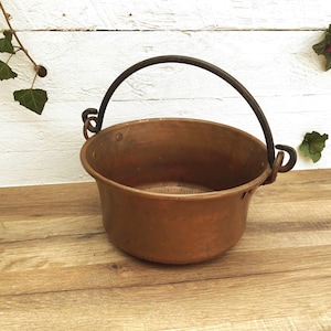 May include: A copper cauldron with a dark metal handle. The pot has a rounded shape and is designed with two small loops on either side for hanging. The copper has a warm, reddish-brown hue.