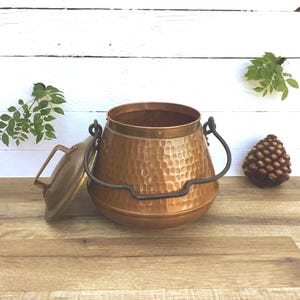 May include: A copper cauldron with a hammered texture, a brass rim, and a hinged lid. The cauldron has a black handle and is set on a wooden surface. A pinecone and greenery are in the background.