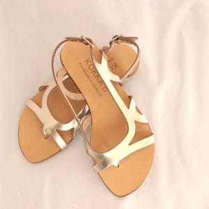 May include: Gold leather sandals with a strappy design and a buckle closure. The sandals are handmade in Greece and have the brand name "KOKKA" printed on the strap.
