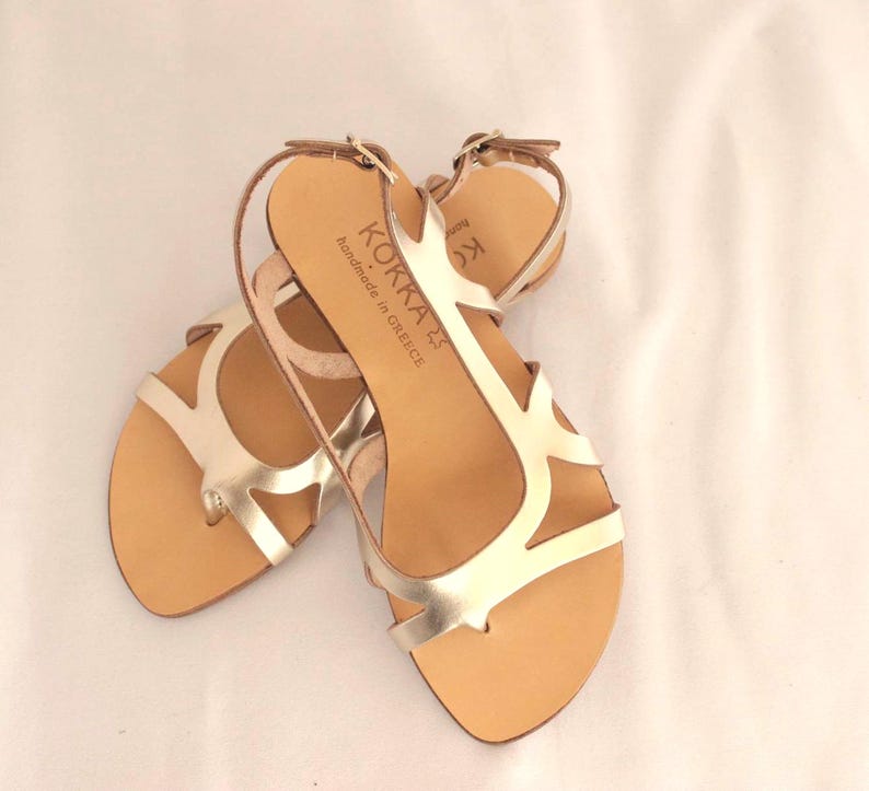 May include: Gold leather sandals with a strappy design and a buckle closure. The sandals are handmade in Greece and have the brand name "KOKKA" printed on the strap.