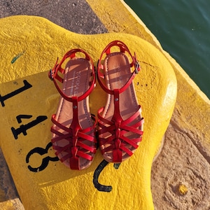 Two red sandals with a criss-cross strap design. The sandals have a brown leather insole and are sitting on a yellow surface.