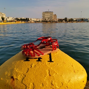 Puede incluir: Un par de sandalias de cuero rojo con un dise&ntilde;o de tiras cruzadas. Las sandalias est&aacute;n sobre una boya amarilla en un puerto.