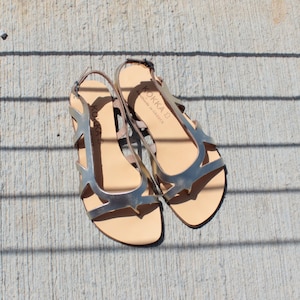 May include: A pair of silver leather sandals with an open toe and ankle strap. The sandals have a cut-out design and the brand name "KOKKA" is printed on the straps. "Handmade in Greece" is also printed on the straps.
