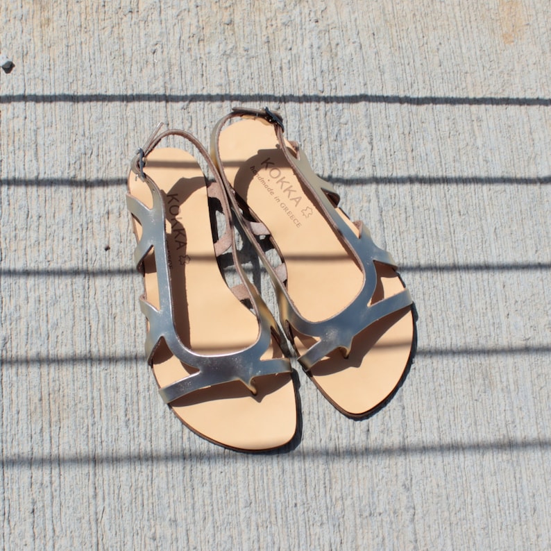 May include: A pair of silver leather sandals with an open toe and ankle strap. The sandals have a cut-out design and the brand name "KOKKA" is printed on the straps. "Handmade in Greece" is also printed on the straps.