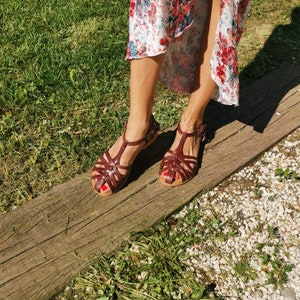 Puede incluir: Un par de sandalias de cuero marr&oacute;n con un dise&ntilde;o de tiras cruzadas. Las sandalias las lleva una persona de pie sobre una superficie de madera.