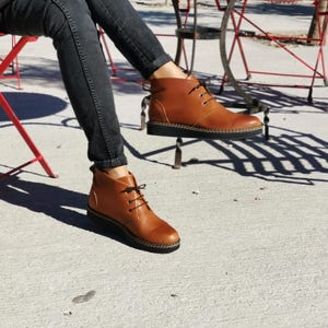 Puede incluir: Botines de cuero marrón con suelas y cordones negros. Las botas tienen un diseño cosido alrededor de la suela. La persona lleva vaqueros negros. Las botas se muestran al aire libre.