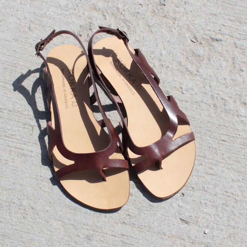 May include: A pair of brown leather sandals with a criss-cross strap design. The sandals have a thong toe and an ankle strap. The sandals are handmade in Greece and have the brand name "KOKKA" printed on the insole.