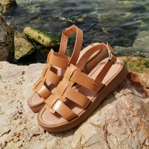 May include: A pair of brown leather sandals with a thick platform sole and a woven design. The sandals are sitting on a rocky surface.