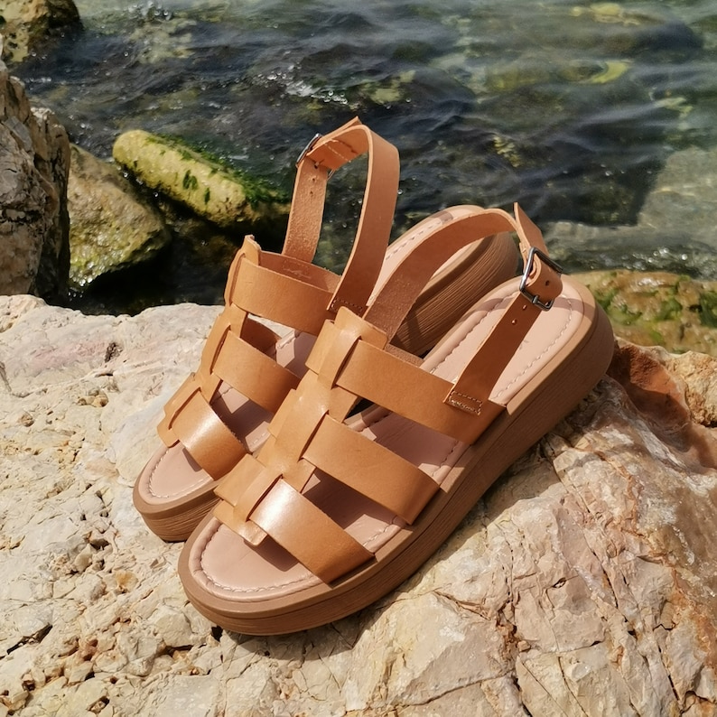 May include: A pair of brown leather sandals with a thick platform sole and a woven design. The sandals are sitting on a rocky surface.
