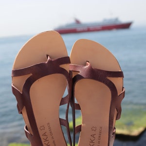 May include: A pair of brown leather sandals with a criss-cross strap design. The sandals have a simple, minimalist style and are made in Greece. The text "KKA made in Greece" is printed on the sole of the sandals.