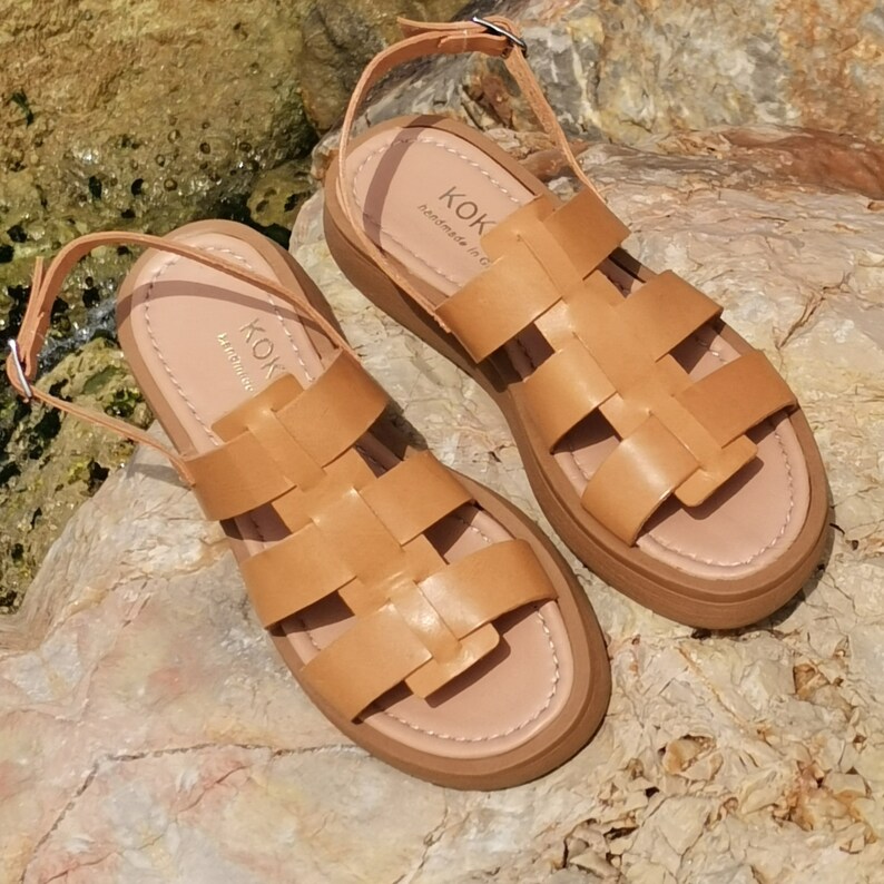 May include: A pair of tan leather sandals with a woven design. The sandals have a thick platform sole and an ankle strap with a buckle closure. The sandals are handmade in Greece and have the brand name "KOK" on the insole.