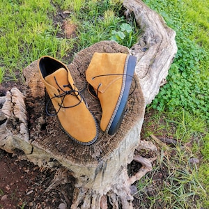 May include: Two brown leather ankle boots with black laces and black soles. The boots are sitting on a tree stump with green grass and brown dirt in the background.