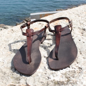 Peut inclure: Une paire de sandales en cuir marron avec une boucle de fermeture. Les sandales sont sur une surface en pierre de couleur claire.
